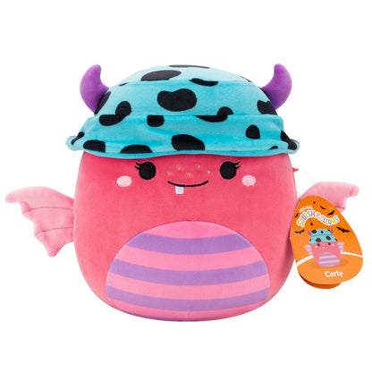 The pink Carly Squishmallow monster plush features purple horns poking through a blue spotted cap, small pink wings, and a striped belly