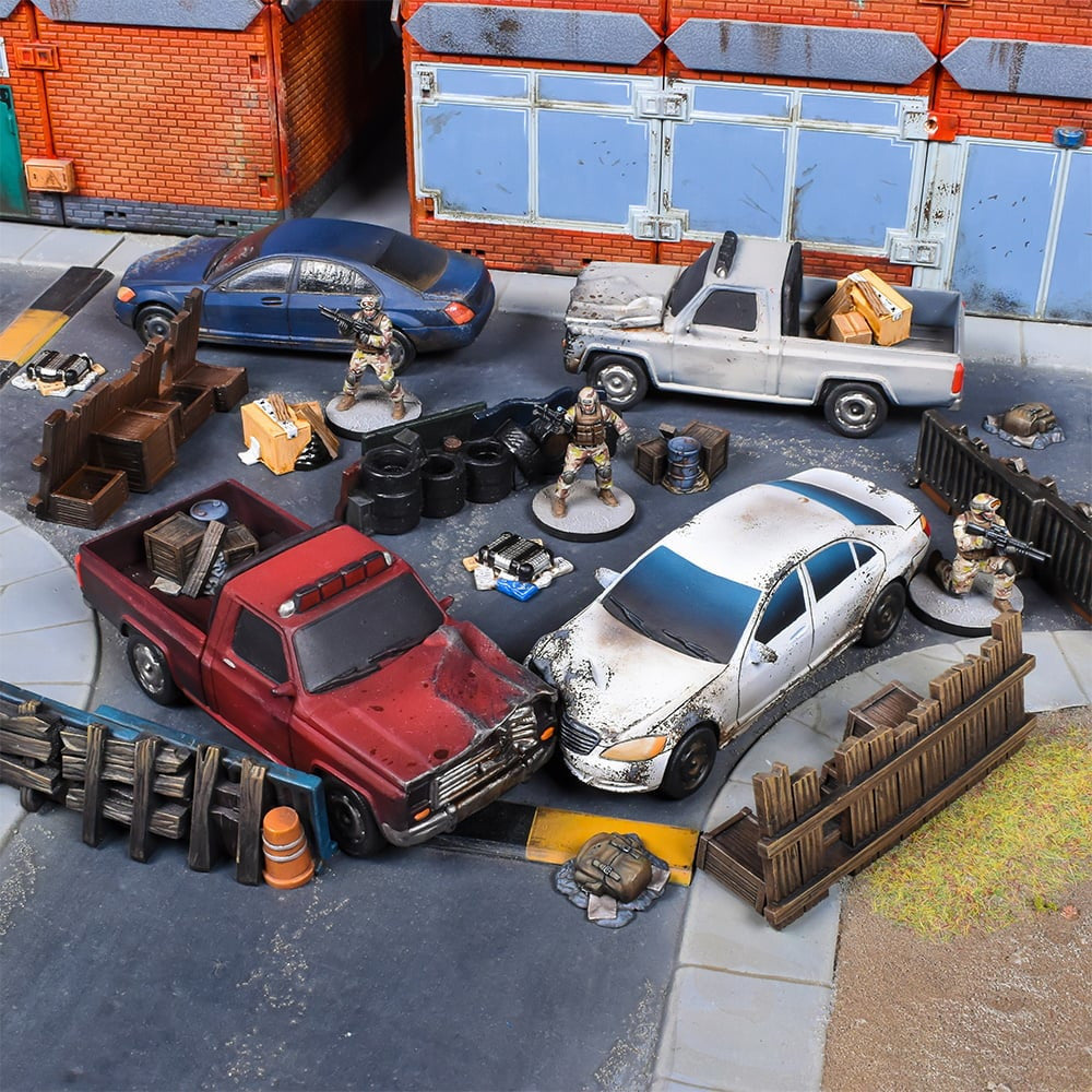 Miniature urban wargame setup with damaged cars, armed soldiers, wooden barricades, and debris on a street, flanked by brick buildings