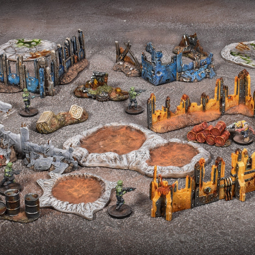 Collection of sci-fi wargaming terrain featuring ruined blue, orange, and grey buildings, impact craters, barrels, and green-clad miniature soldiers