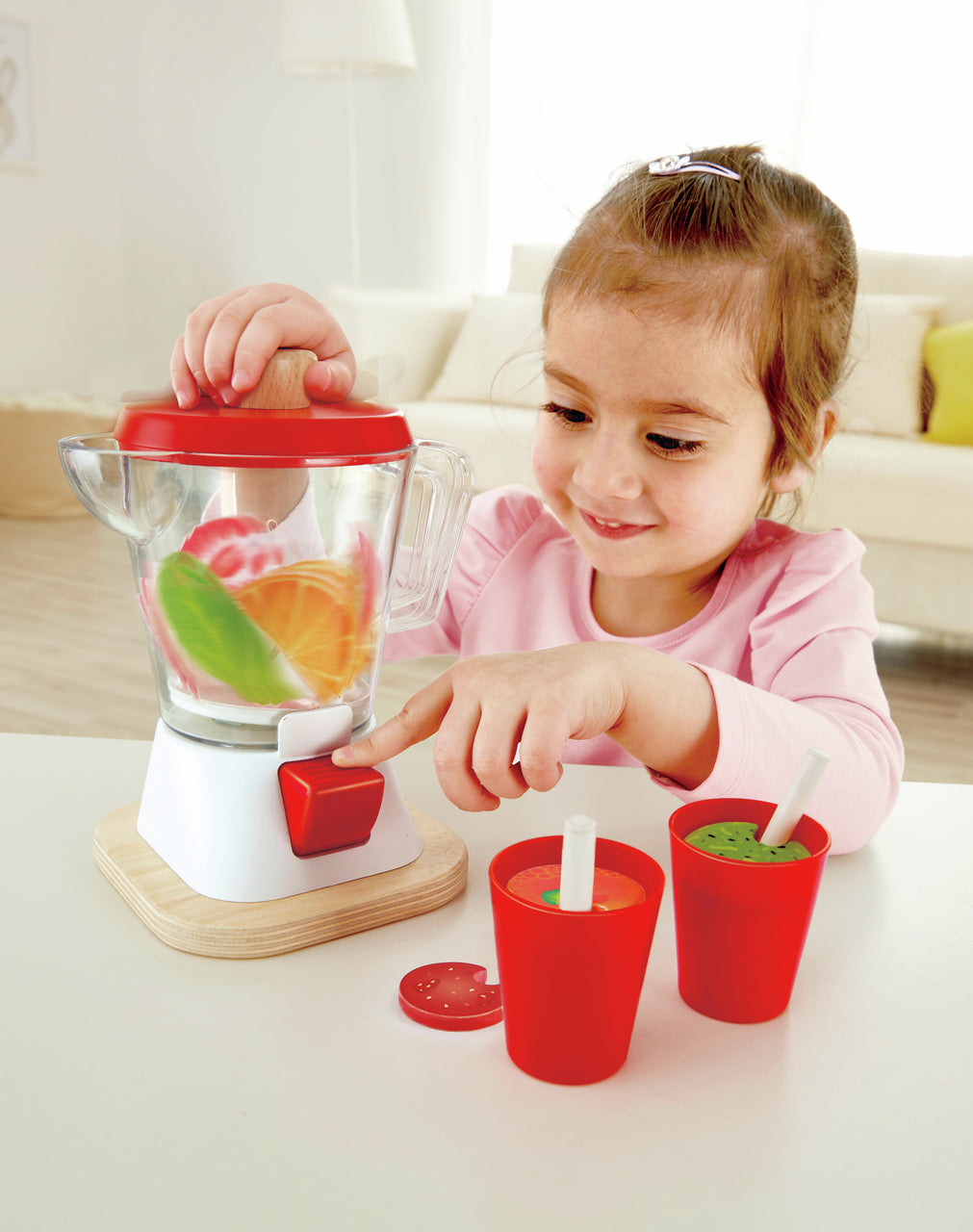 Smiling young girl uses a red and white toy blender set, pressing the button to mix colorful fruit slices