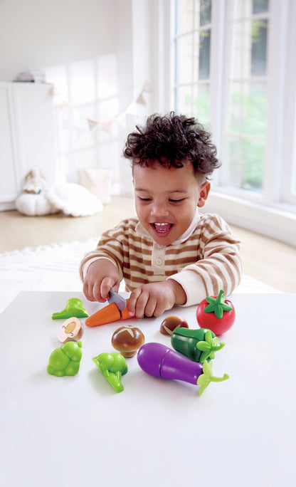 Smiling child cutting a toy carrot with a knife, surrounded by colorful play food