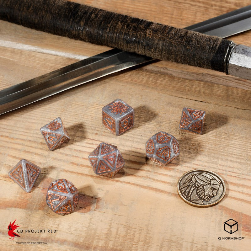 Seven ornate silver and copper polyhedral dice and a coin on a wooden table, next to a sword