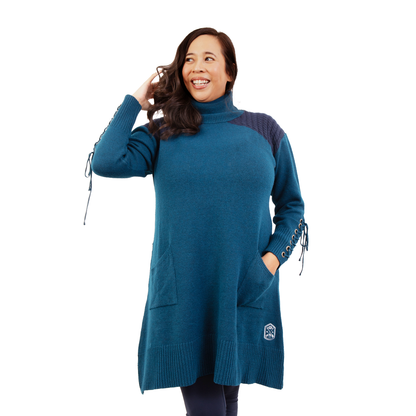 Smiling woman in a teal turtleneck sweater dress with laced sleeves, hands in hair and pocket