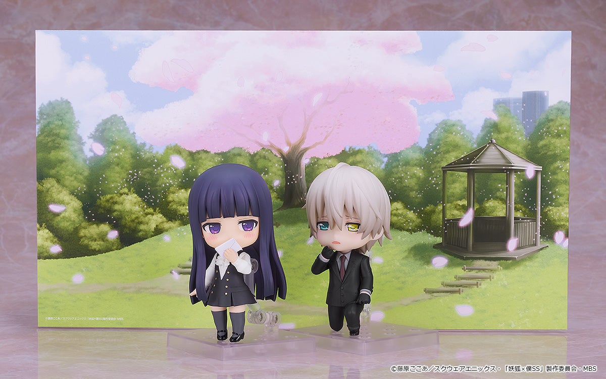 Two chibi-style anime figures in a spring park. A girl holds an envelope, a boy in a suit looks thoughtful.
