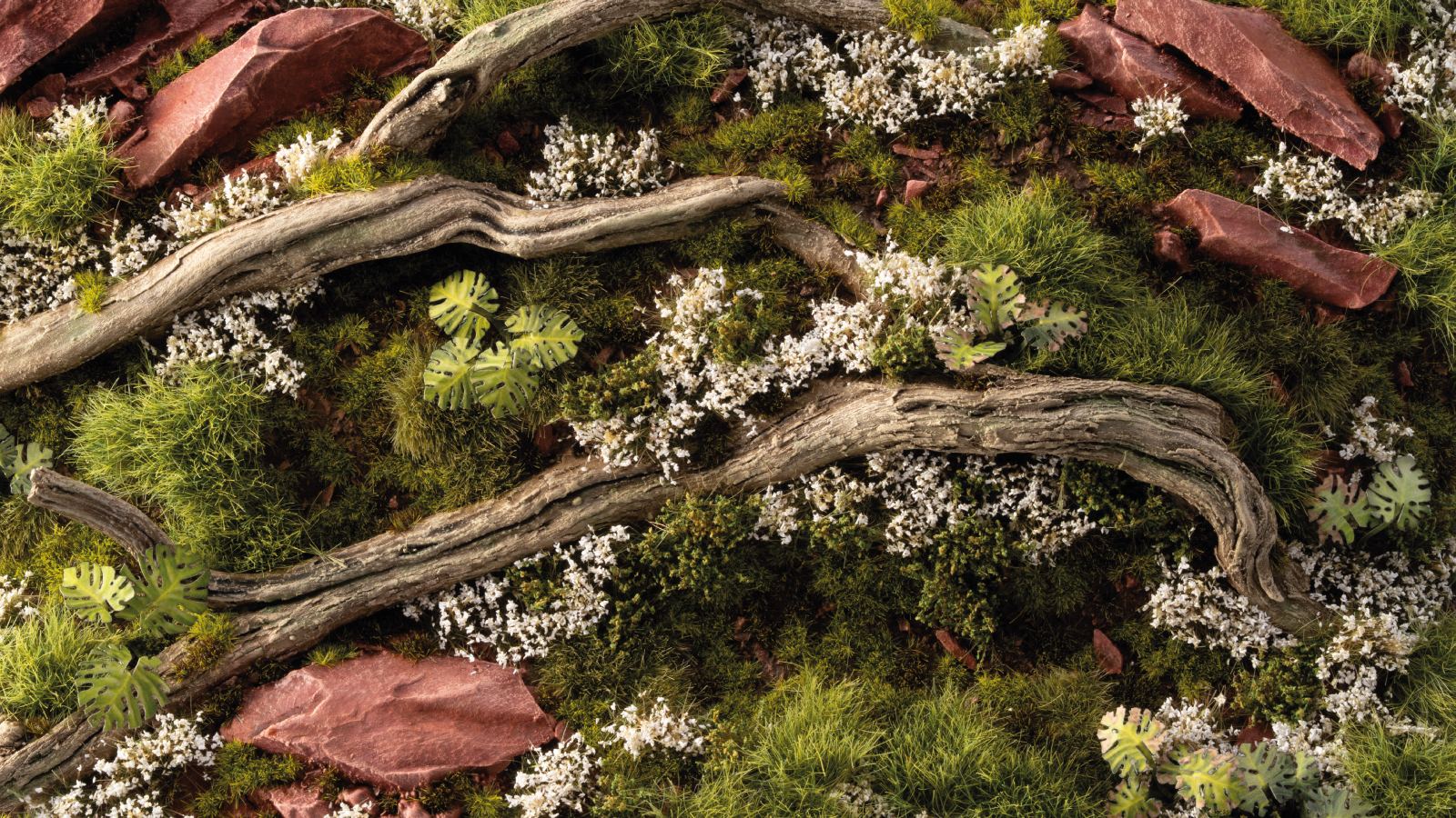 Detailed model terrain featuring twisted deadwood, lush green moss and grass, reddish rocks, and small white flowers