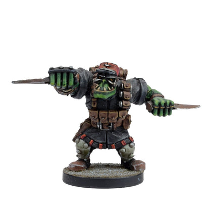 A miniature of a green orc warrior with tusks, wearing a red cap and tactical vest, holding a knife in each hand