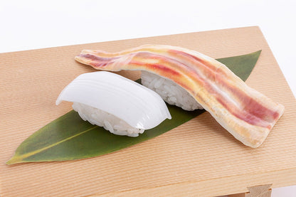 Two sushi pieces: one with white squid-like topping, one with a marbled bacon-like topping, on a leaf on a wooden board